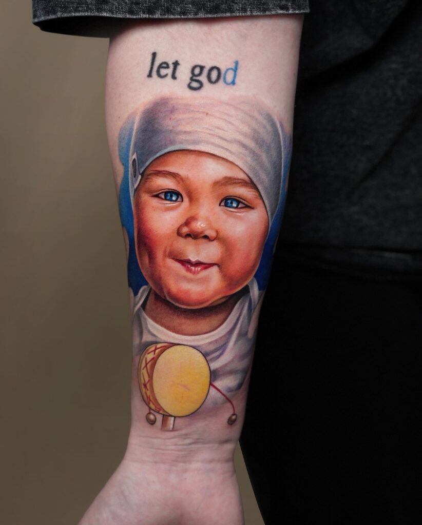 Realistic color portrait tattoo of a smiling baby holding a toy drum on a forearm sleeve